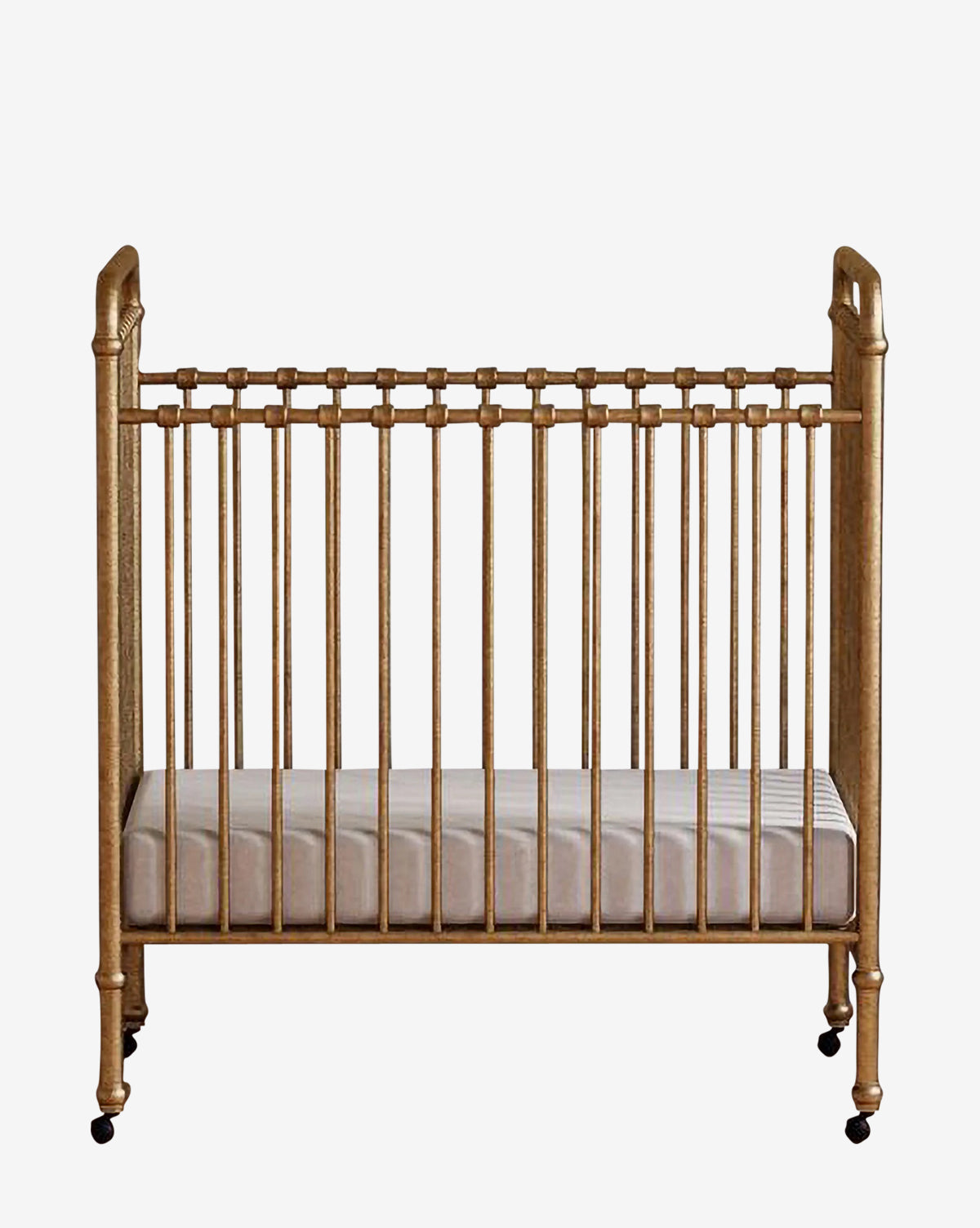 The Abigail Mini Crib by Million Dollar Baby is a gold-colored vintage crib featuring vertical slats, decorative rounded corners, a white mattress, and small black wheels, displayed against a plain white background. Vendixs