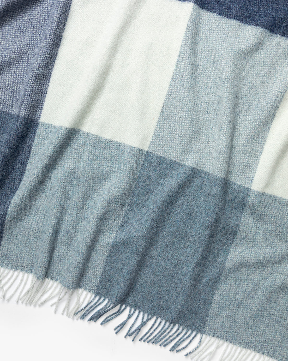 The Abner Throw by Fibre is a soft blue plaid blanket with a large blue, gray, and white pattern, finished with fringed edges and crafted from recycled alpaca wool for an eco-friendly touch. Vendixs