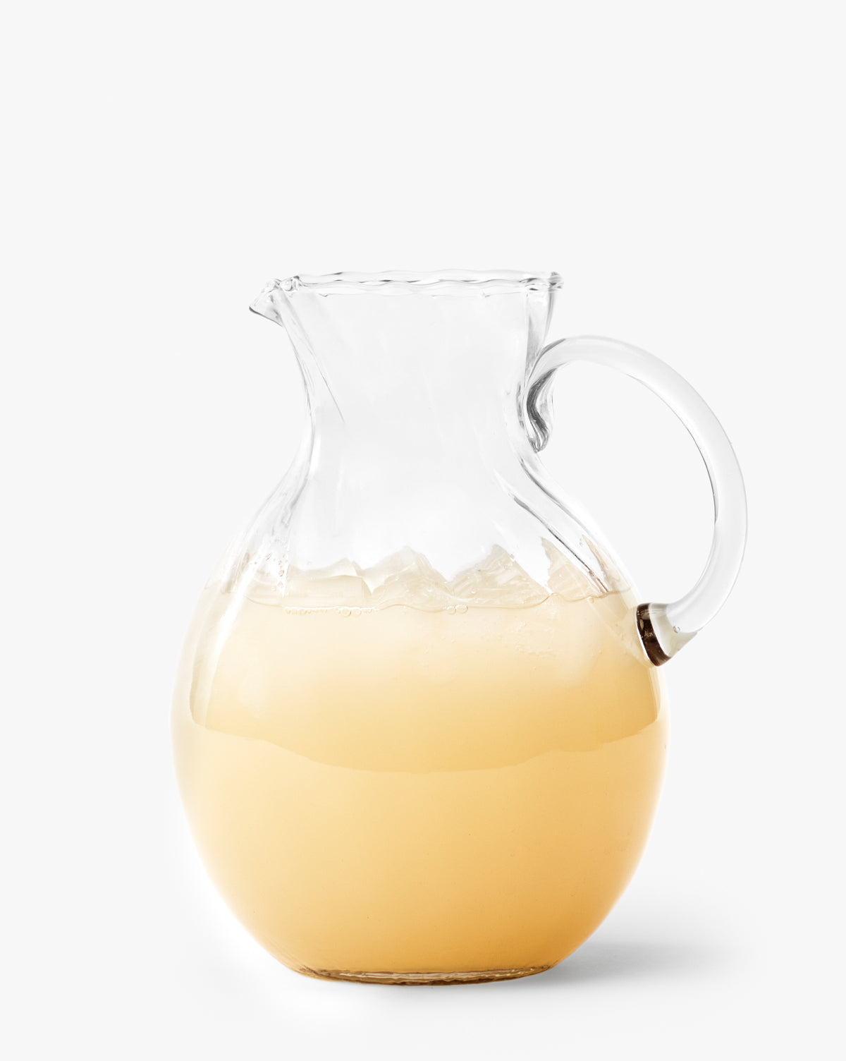 The Adderley Pitcher, a mouth-blown glass piece with a scalloped rim, holds light yellow juice and ice cubes, displayed against a plain white background by McGee & Co.