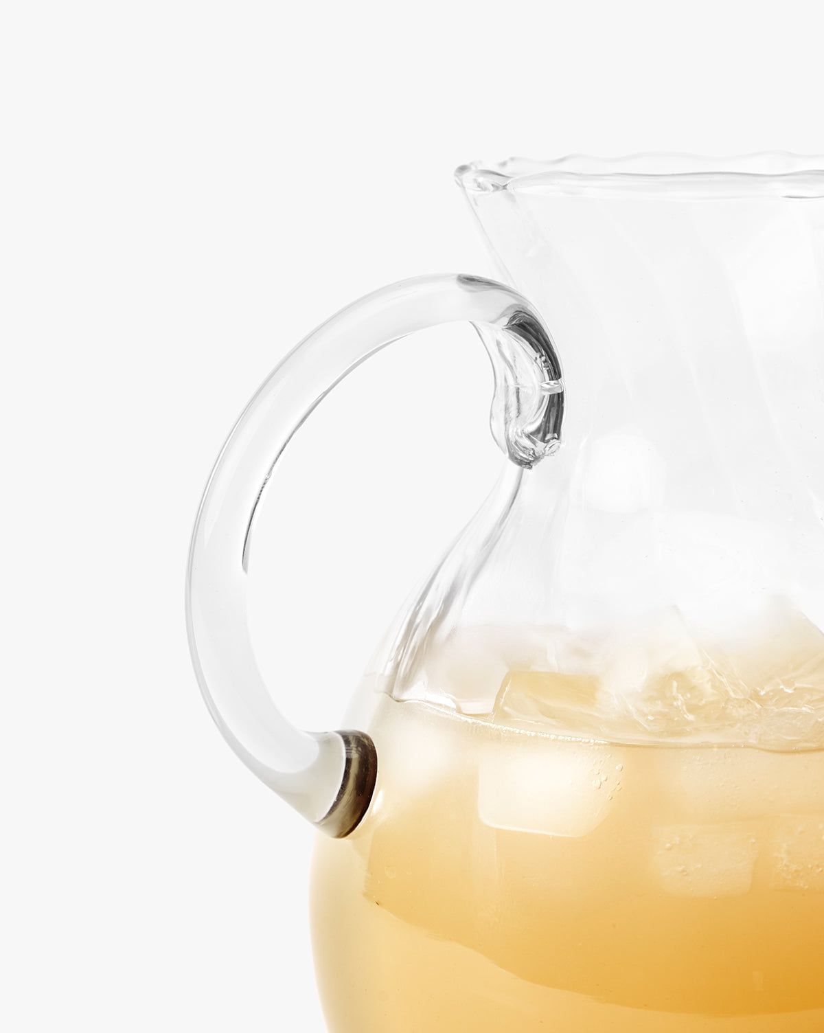 A close-up of the Adderley Pitcher, a mouth-blown glass piece with a scalloped rim, filled with an iced light-colored drink. The focus is on the pitcher’s handle and ice, set against a plain white background, by McGee & Co.