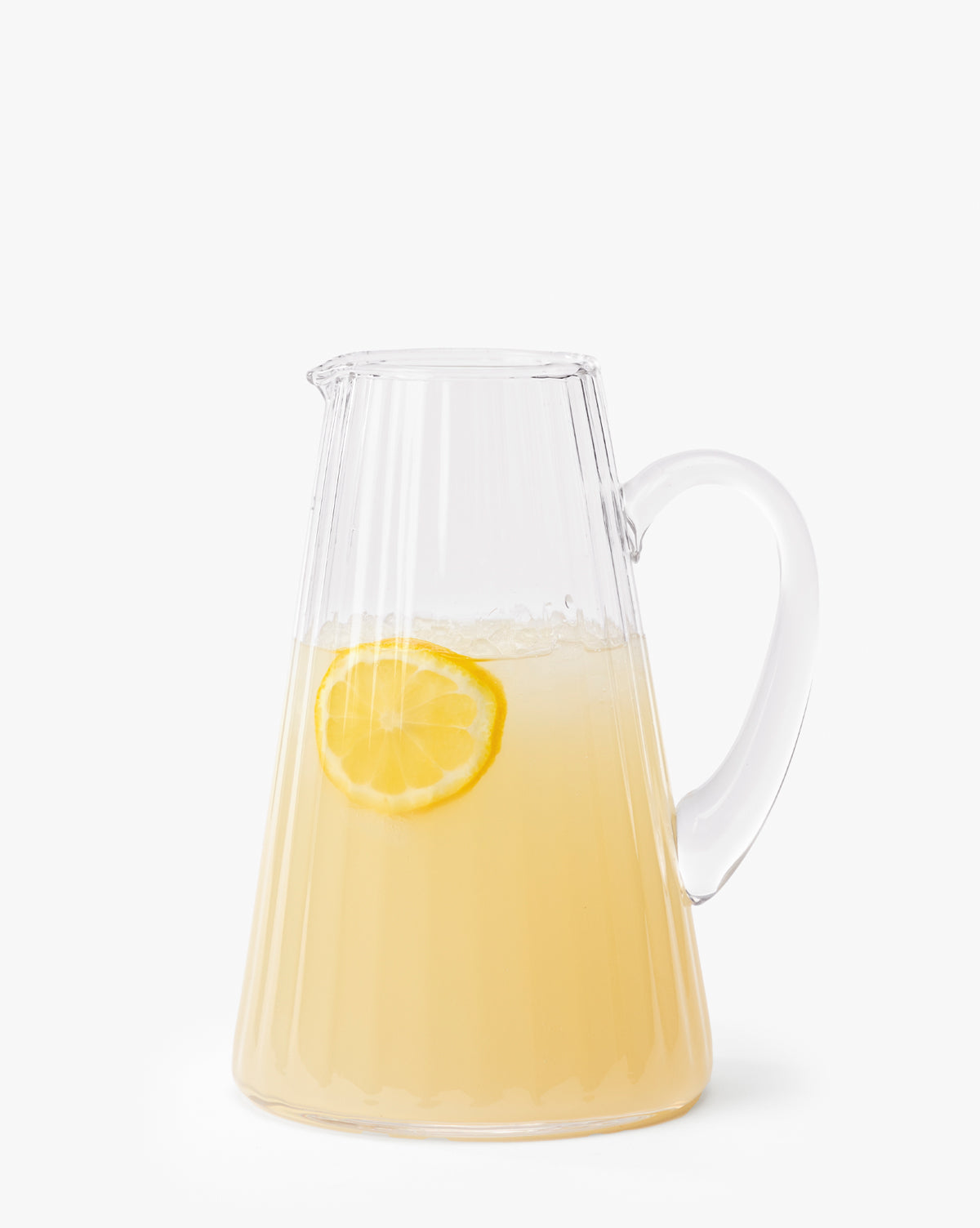The Adonis Ribbed Glass Pitcher, filled with pale yellow lemonade and a lemon slice floating inside, sits against a plain white background—ideal for effortless kitchen styling, by Vendixs