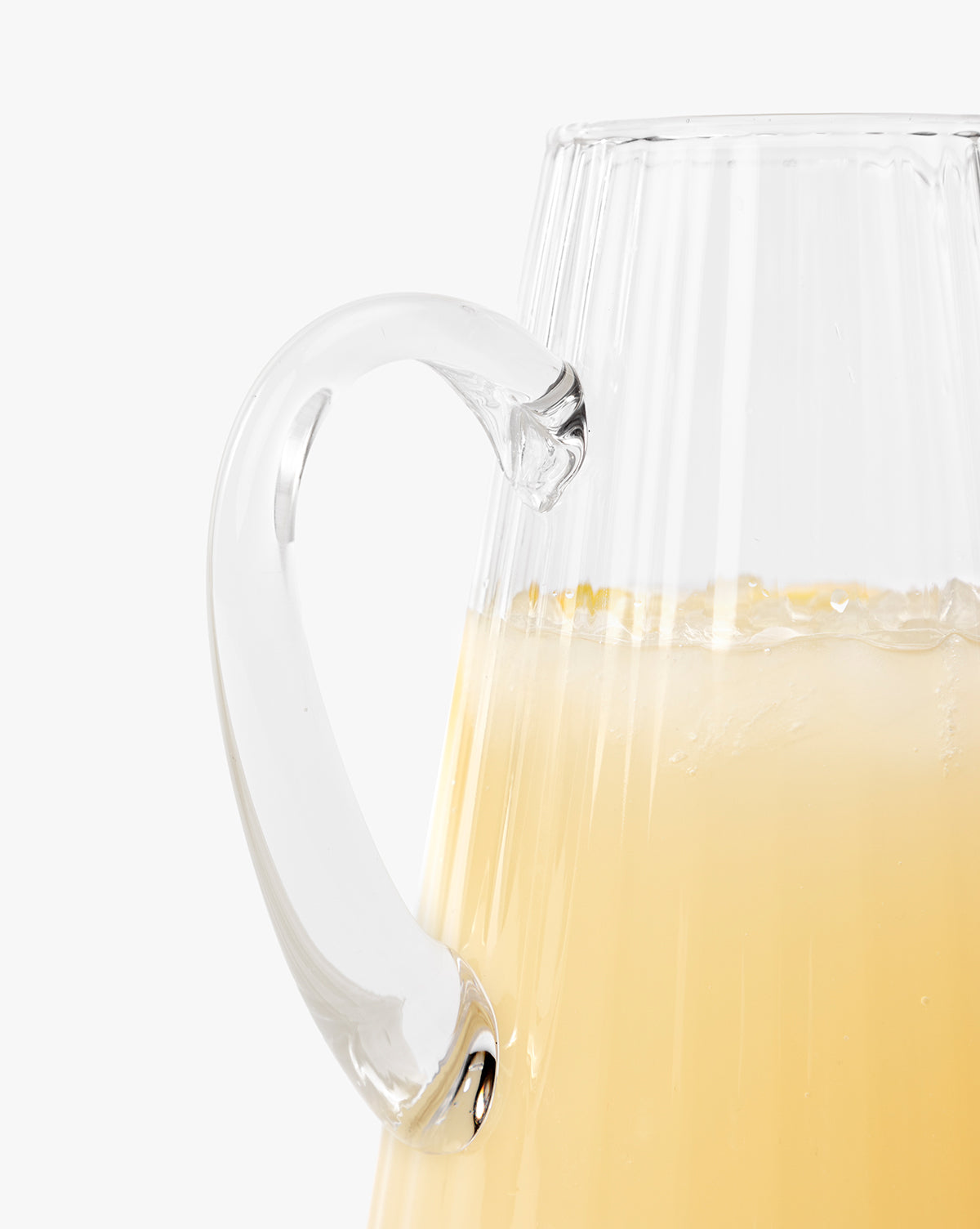 Close-up of the Adonis Ribbed Glass Pitcher filled with a pale yellow drink and ice, showcasing its textured surface and curved handle—ideal for stylish kitchen décor against a white backdrop, by McGee & Co.