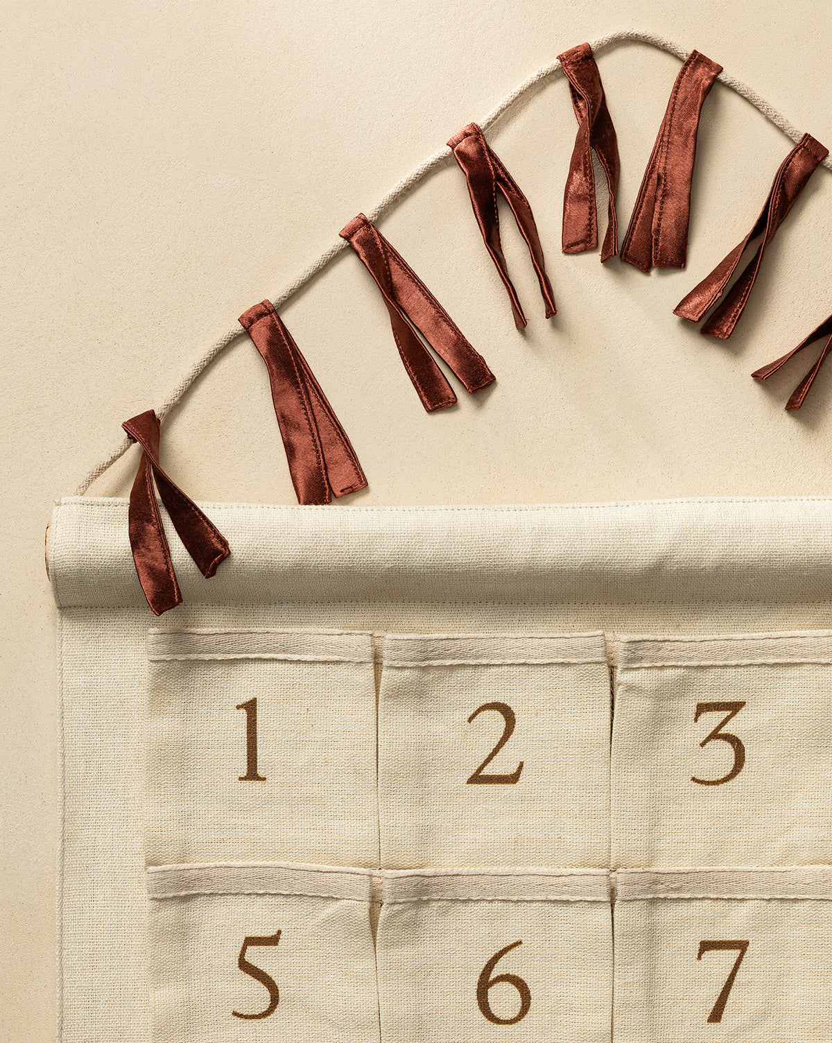 The Advent Calendar is a cotton-linen wall hanging in brown with numbered pockets (1-6 visible), tied to a string with red ribbons. It’s perfect for starting a holiday tradition against a neutral beige background, by Vendixs