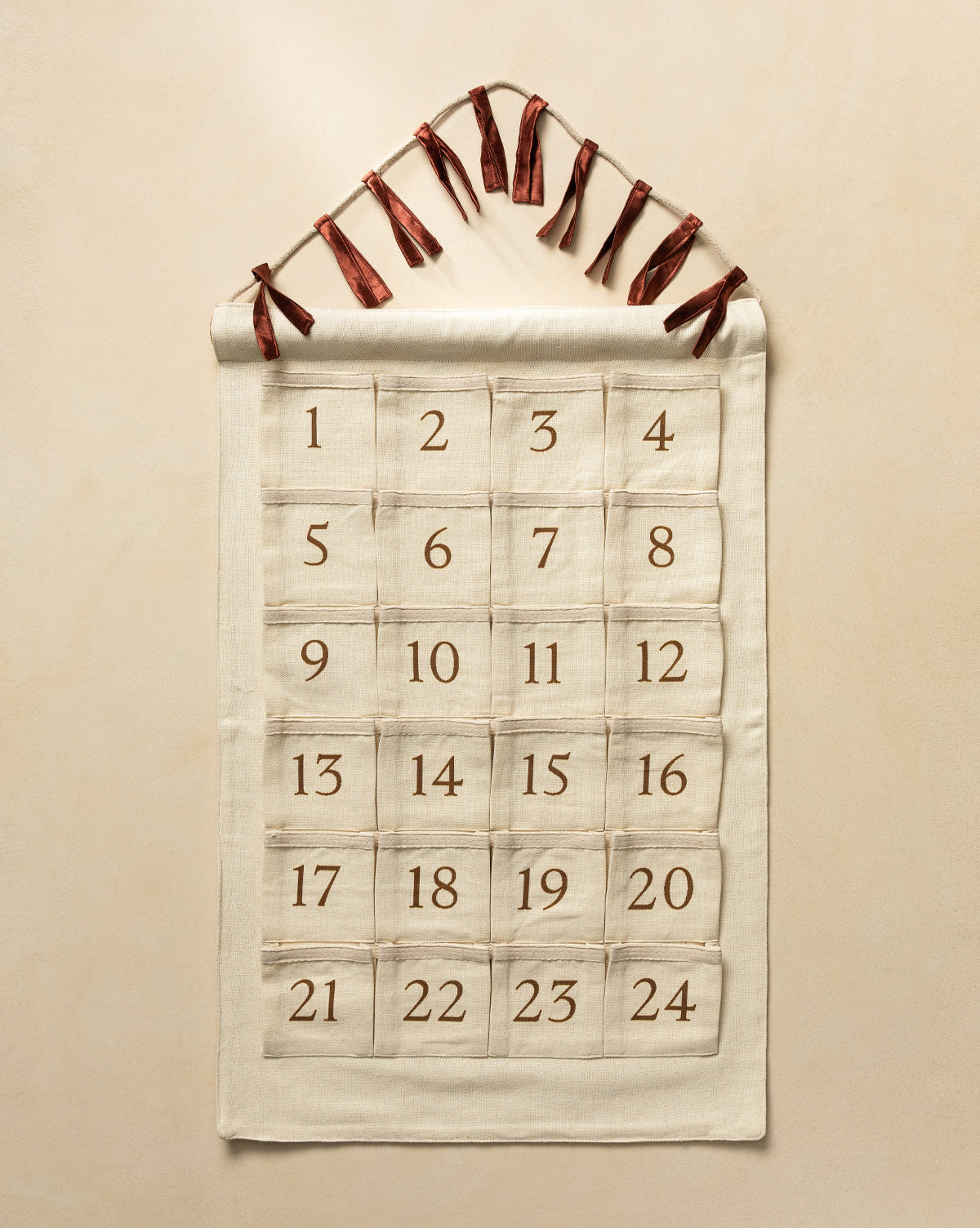The Advent Calendar is a cream-colored cotton linen piece with 24 numbered pockets in a grid, hanging from a rod with brown ribbons—a charming way to count down to Christmas, by Vendixs