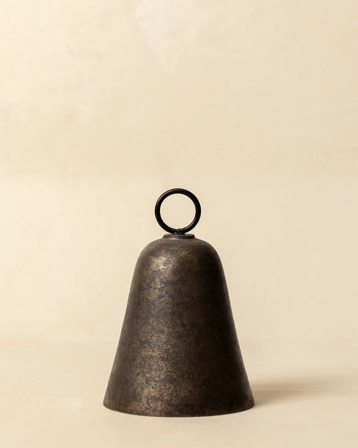 The Aged Brass Bell, featuring a dark finish and round top handle, rests on a light beige surface, bringing vintage charm to the simple background, by Vendixs
