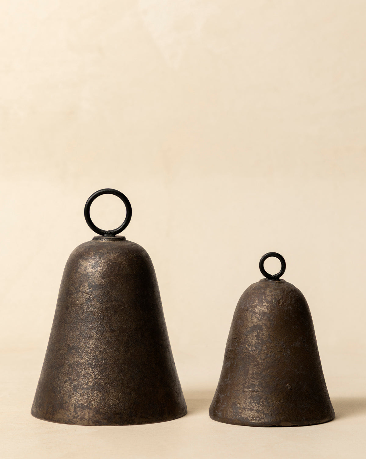 Two Aged Brass Bells with round black handles sit side by side on a light beige surface. The larger and taller bell pairs with a smaller, shorter one, both displaying vintage charm, by Vendixs