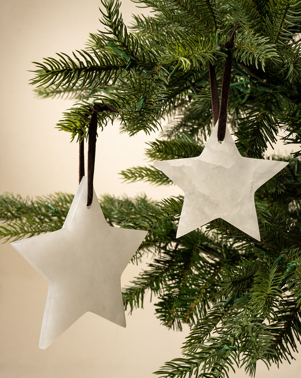 The Alabaster Star Ornaments (Set of 2) hang from brown ribbons on a green Christmas tree, bringing elegant style to your festive décor against a neutral beige background, by Vendixs