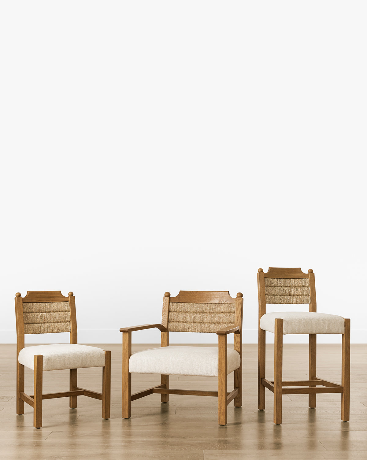 Three wooden chairs, featuring the Makerspalm Alberta Counter Stool in honey oak with seagrass-woven backrests and cream cushions, are set on a light wood floor against a white background. The chairs differ in armrest style and height. Vendixs