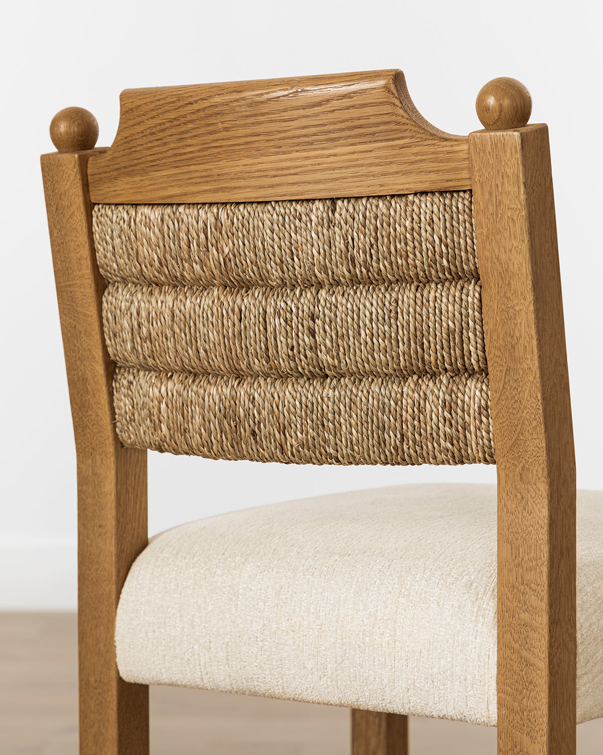 The Makerspalm Alberta Counter Stool features decorative ball finials, a woven seagrass backrest, and a cushioned beige fabric seat. It’s pictured on a light-colored floor against a white wall. Vendixs
