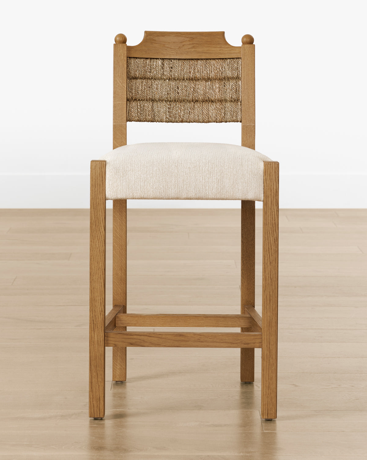 The Makerspalm Alberta Counter Stool has a woven seagrass backrest, cushioned beige seat, and honey oak frame, shown on a light wood floor with a plain white wall backdrop. Vendixs