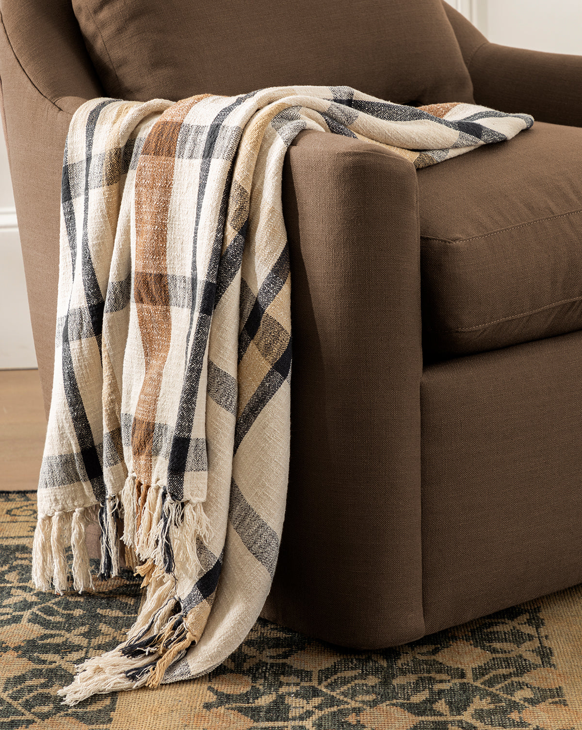 The Raj Albright Plaid Throw, featuring beige, brown, and black stripes, is draped over a brown upholstered armchair. This cozy cotton blanket adds warmth and style to the chair atop a patterned rug in a warmly lit room by Vendixs