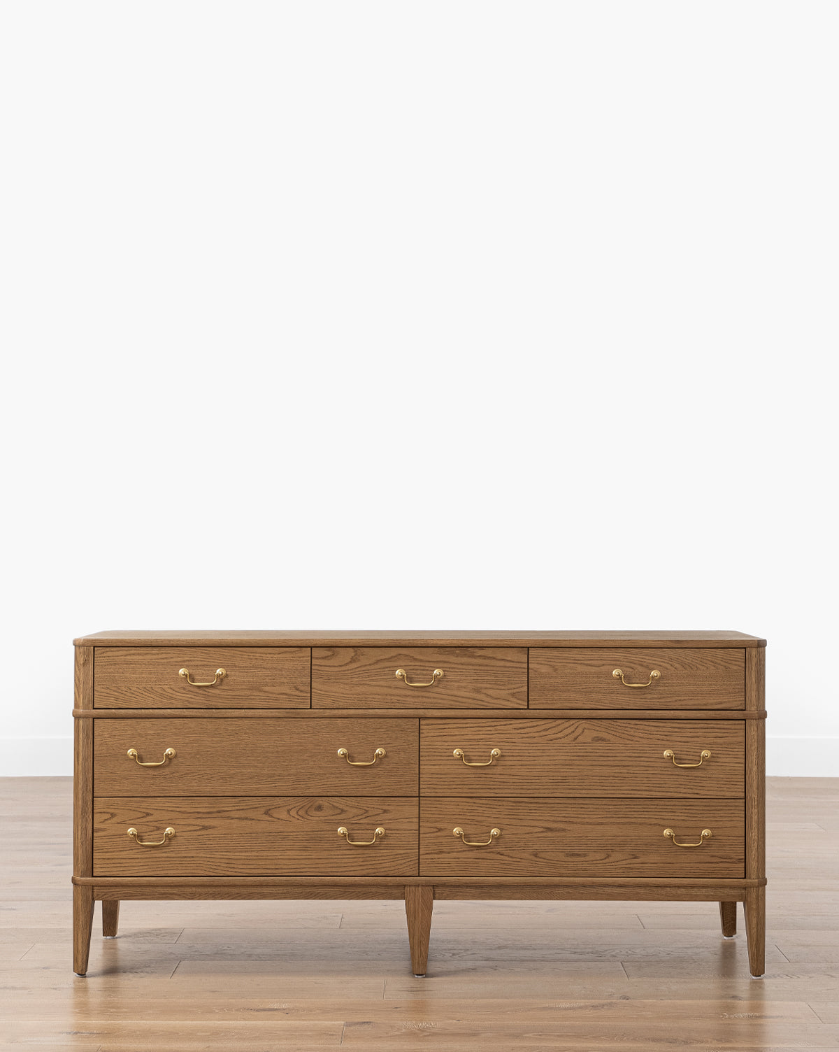 The Makerspalm Alessandro Dresser is a honey oak piece with nine soft-close drawers and brass handles, standing on four tapered legs atop a light wood floor against a plain white background, Vendixs
