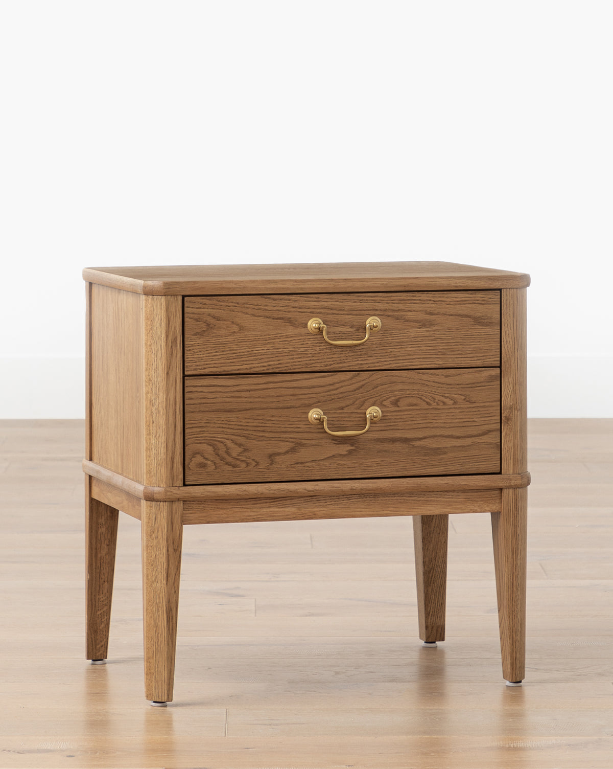 The Alessandra Nightstand, featuring two drawers, brass handles, and tapered legs in elegant honey oak, sits on a light wood floor against a white wall by McGee & Co.