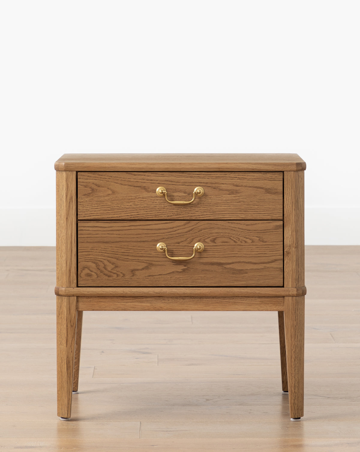 The Alessandra Nightstand by Makerspalm, crafted from solid oak, features two drawers with brass handles and stands on four tapered legs, shown against a light wall and wood floor. Vendixs
