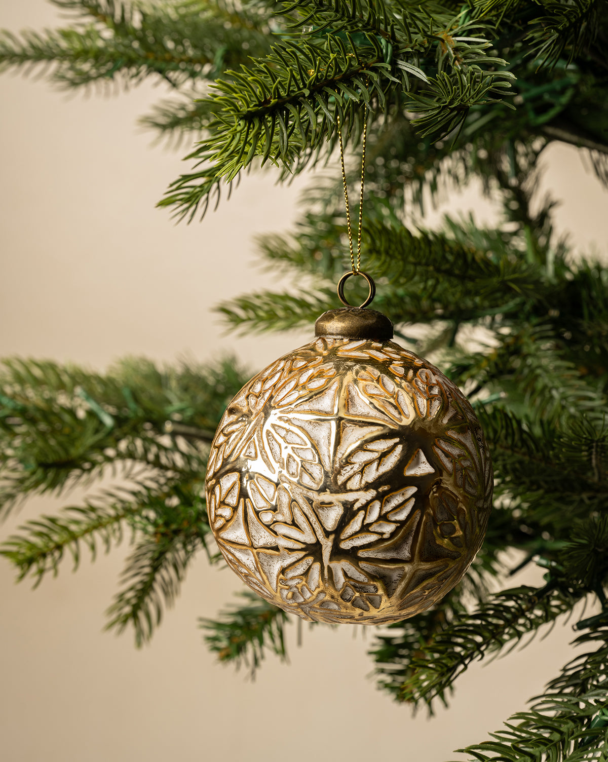 The Alessandro Glass Ornament, featuring an intricate gold pattern, hangs from a green pine branch against a neutral backdrop, bringing timeless charm to your holiday decor, by Vendixs