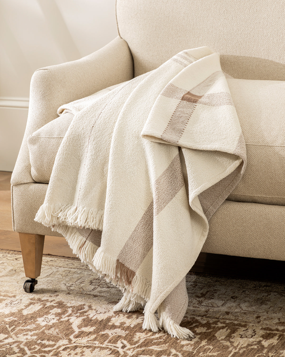 A cream-colored armchair features the ILA HOME FASHIONS Alex Plaid Throw—a beige and tan, fringed blanket—draped over the arm. The chair sits on a patterned rug in a softly lit, cozy room, blending timeless comfort with effortless style from Vendixs