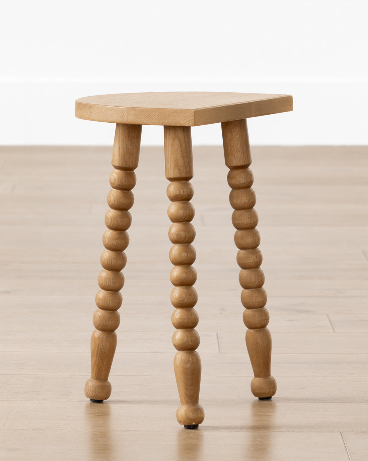 The Alice Honey Wood Stool by Makerspalm features a triangular ash wood seat and spindle legs. Compact and vintage-inspired, it’s perfect as an accent stool for any room. Pictured on a light wood floor against a white background. Vendixs