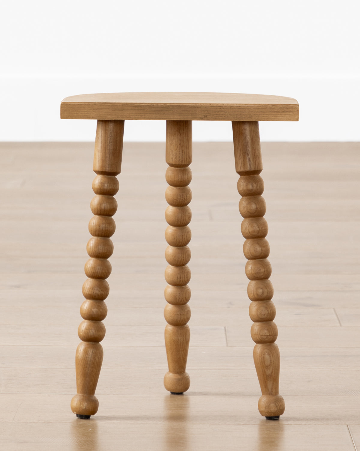 The Alice Honey Wood Stool by Makerspalm features three turned spindle legs and a flat rectangular seat, making it a perfect accent piece. Shown on a light wood floor against a white wall. Vendixs