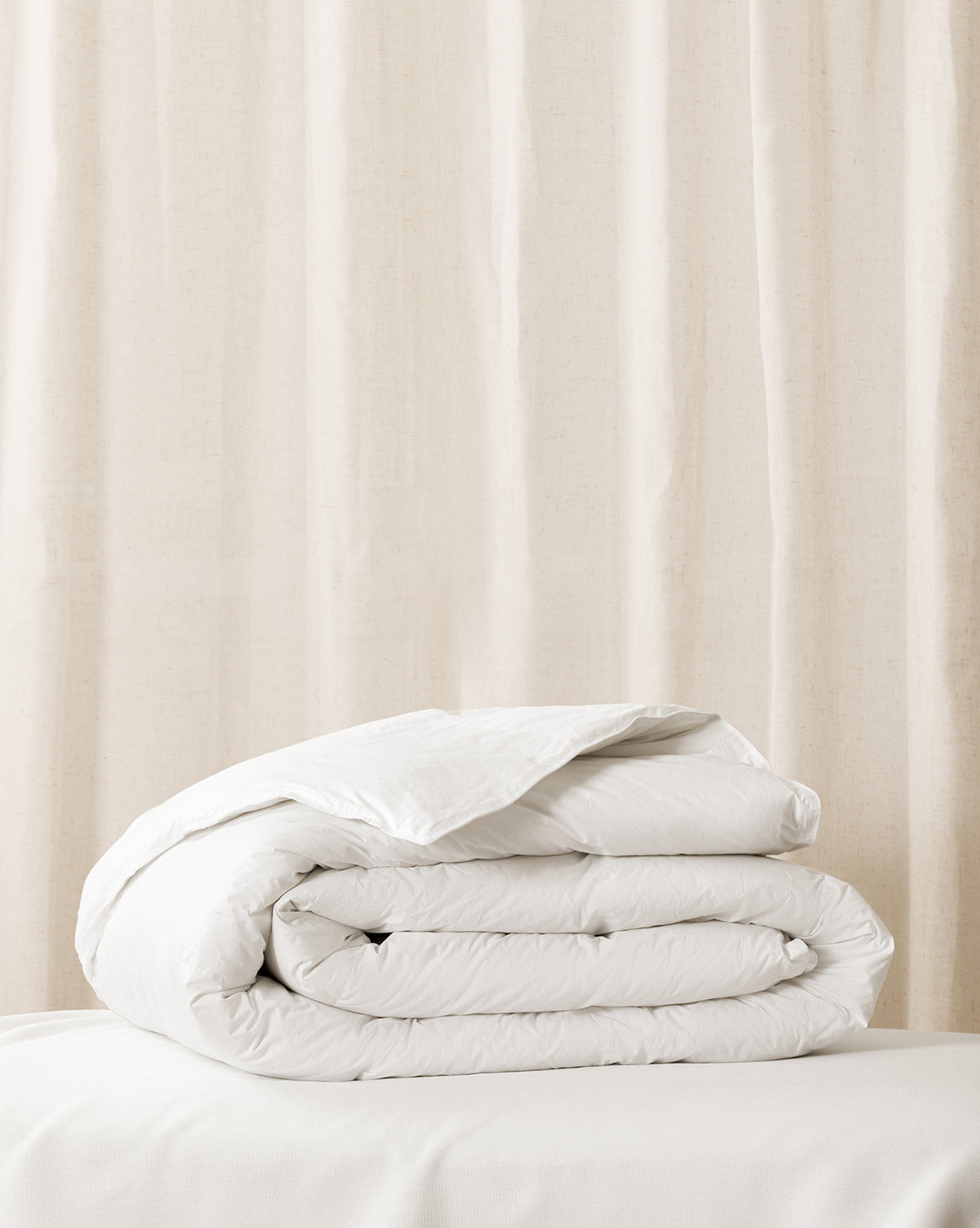 A folded white duvet insert on a bed with a window curtain in the background from Vendixs