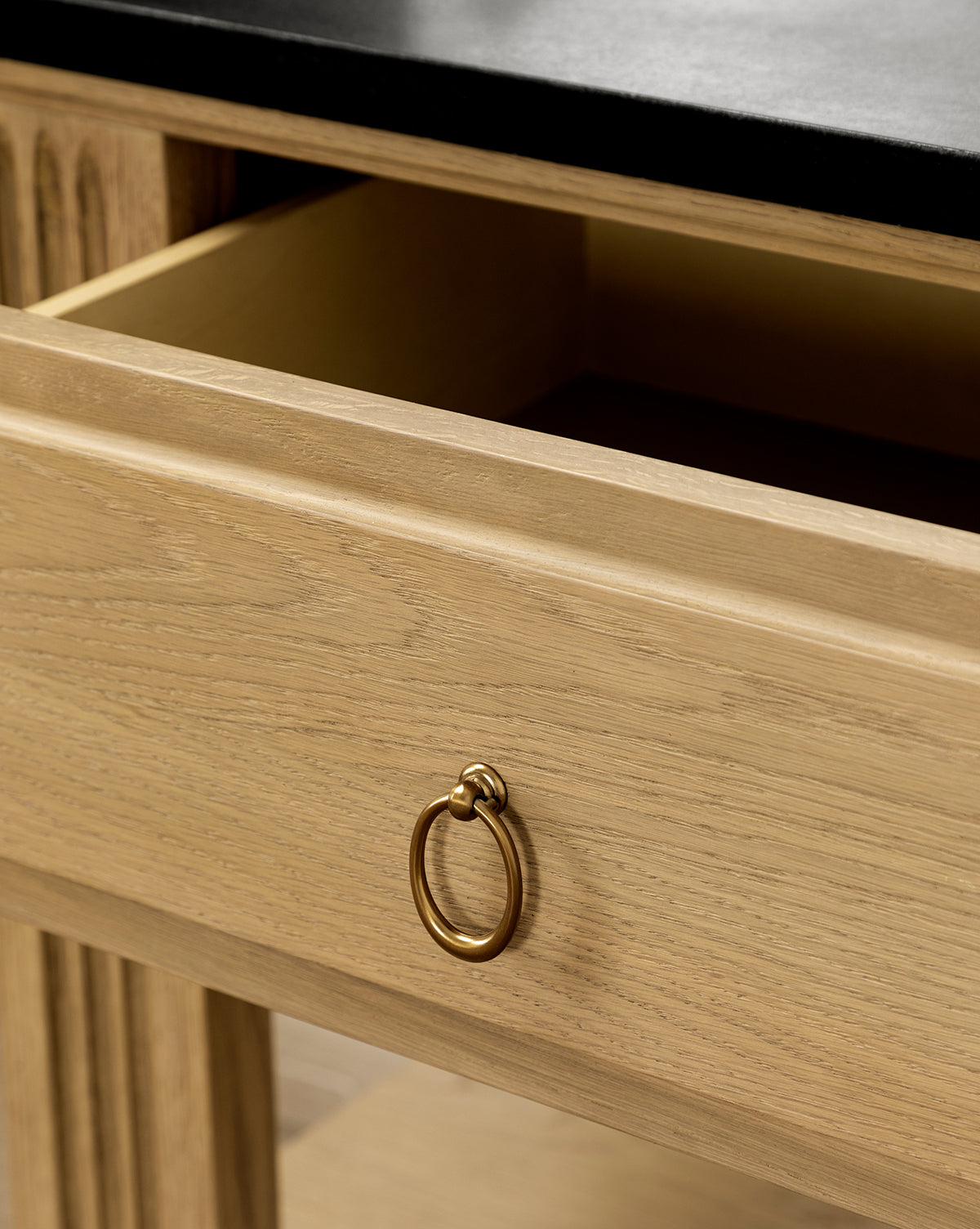 A close-up of the Ambrose Nightstand drawer, featuring a brass ring handle and honed granite top. The light-finished wood and empty interior highlight its elegant, vintage-inspired design for the bedroom, by Vendixs