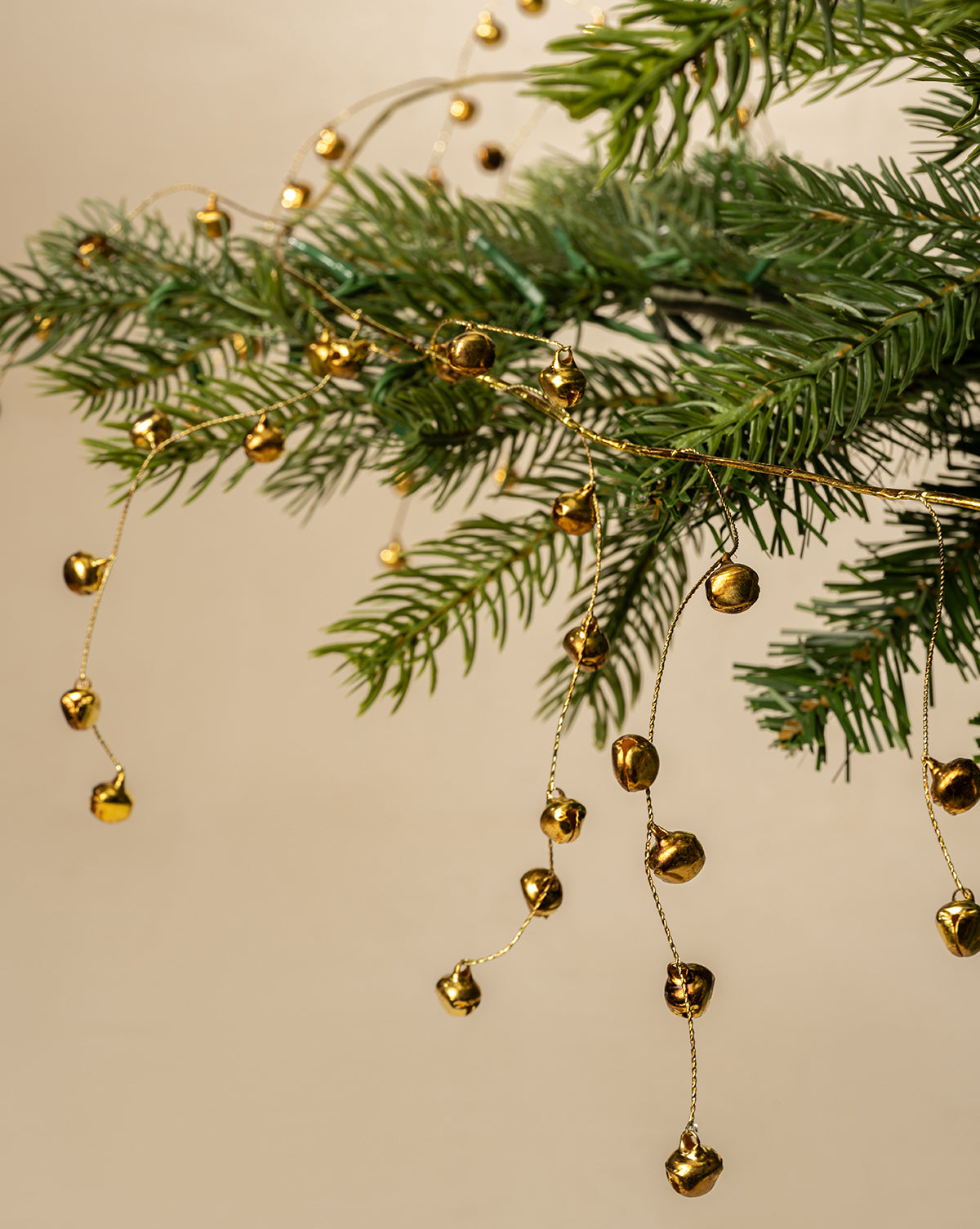 Close-up of evergreen branches decorated with the 6' Angelica Gold Garland, featuring small, shiny bells that add festive holiday charm against a neutral beige background, by Vendixs