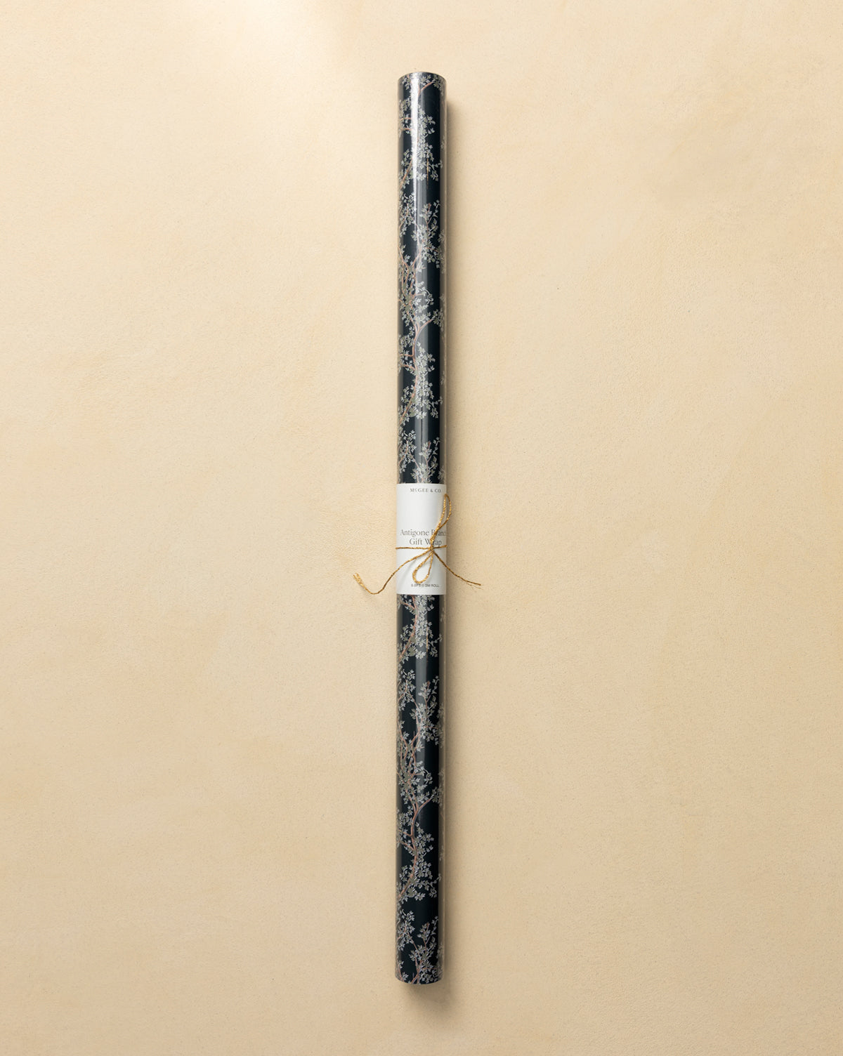 A roll of Antigone Branch Gift Wrap featuring a dark, nature-inspired design with a gold botanical pattern, tied with a thin gold string and labeled, displayed on a light beige background, by Vendixs