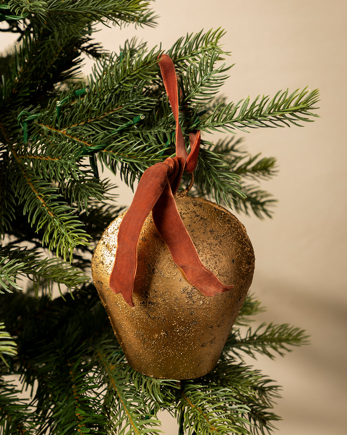 The Antique Gold Metal Bell with Velvet Ribbon hangs from a green Christmas tree branch against a neutral background, adding an elegant touch to your holiday décor, by Vendixs