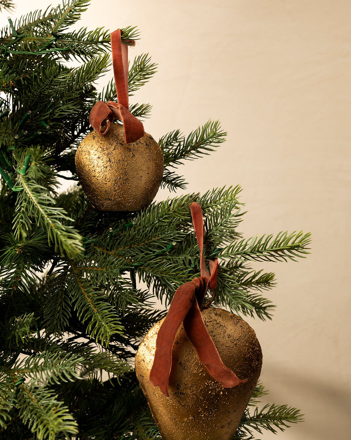 Two Antique Gold Metal Bell with Velvet Ribbon ornaments hang on a green Christmas tree, bringing classic holiday décor to a plain beige background, by Vendixs