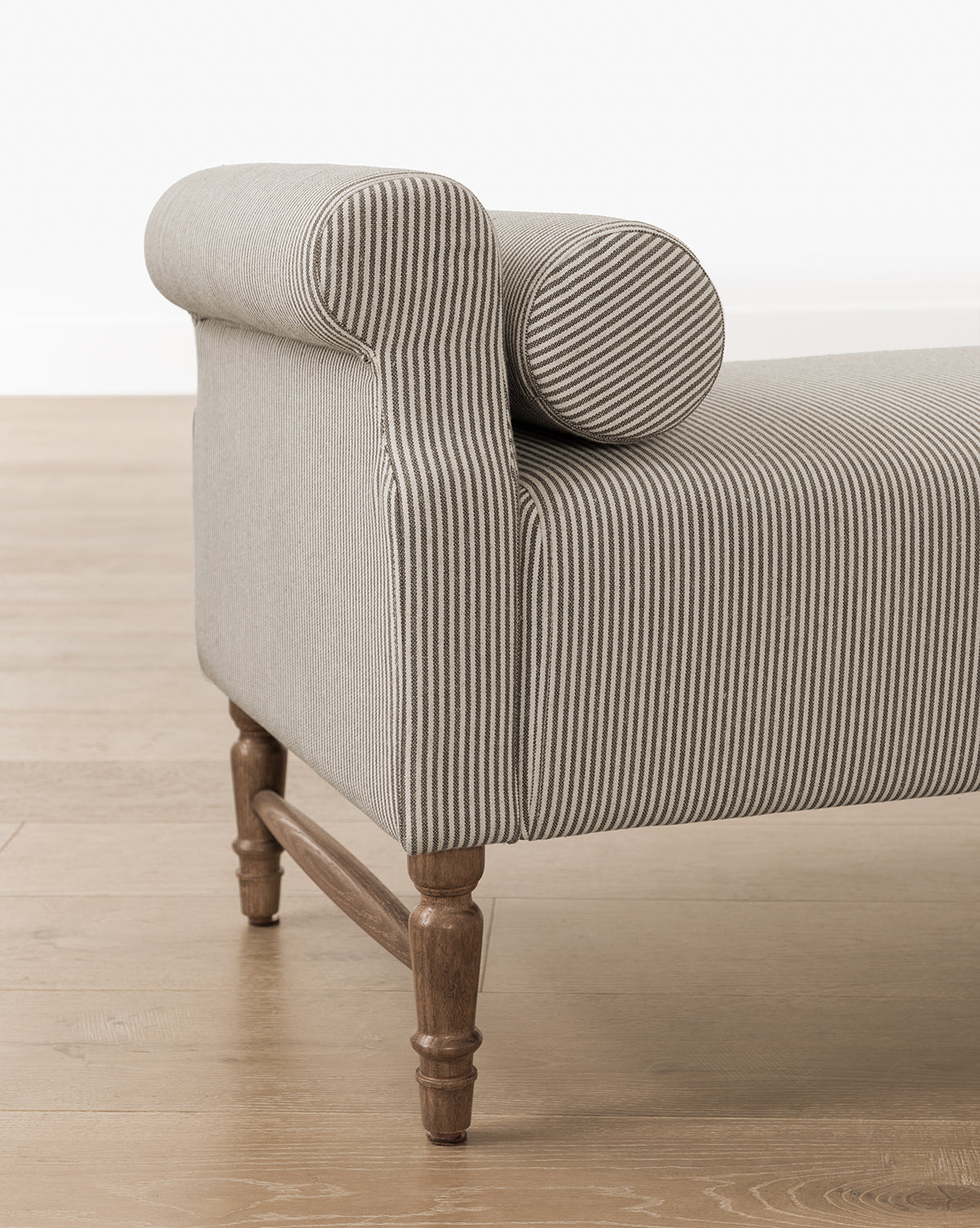 A close-up of the Asher Bench, featuring striped linen upholstery, rolled armrests, matching bolster pillows, and wooden legs set on a light wood floor, by Vendixs