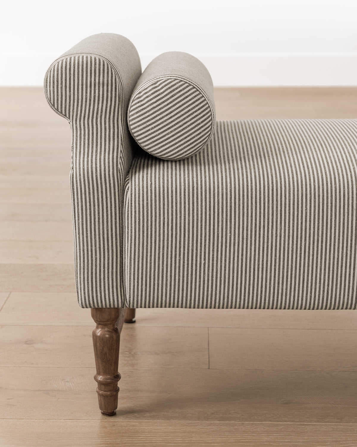 The Asher Bench, featuring gray and white striped linen, a rolled arm with bolster pillows on one end, and wooden legs, sits on a light wood floor against a white wall by Vendixs