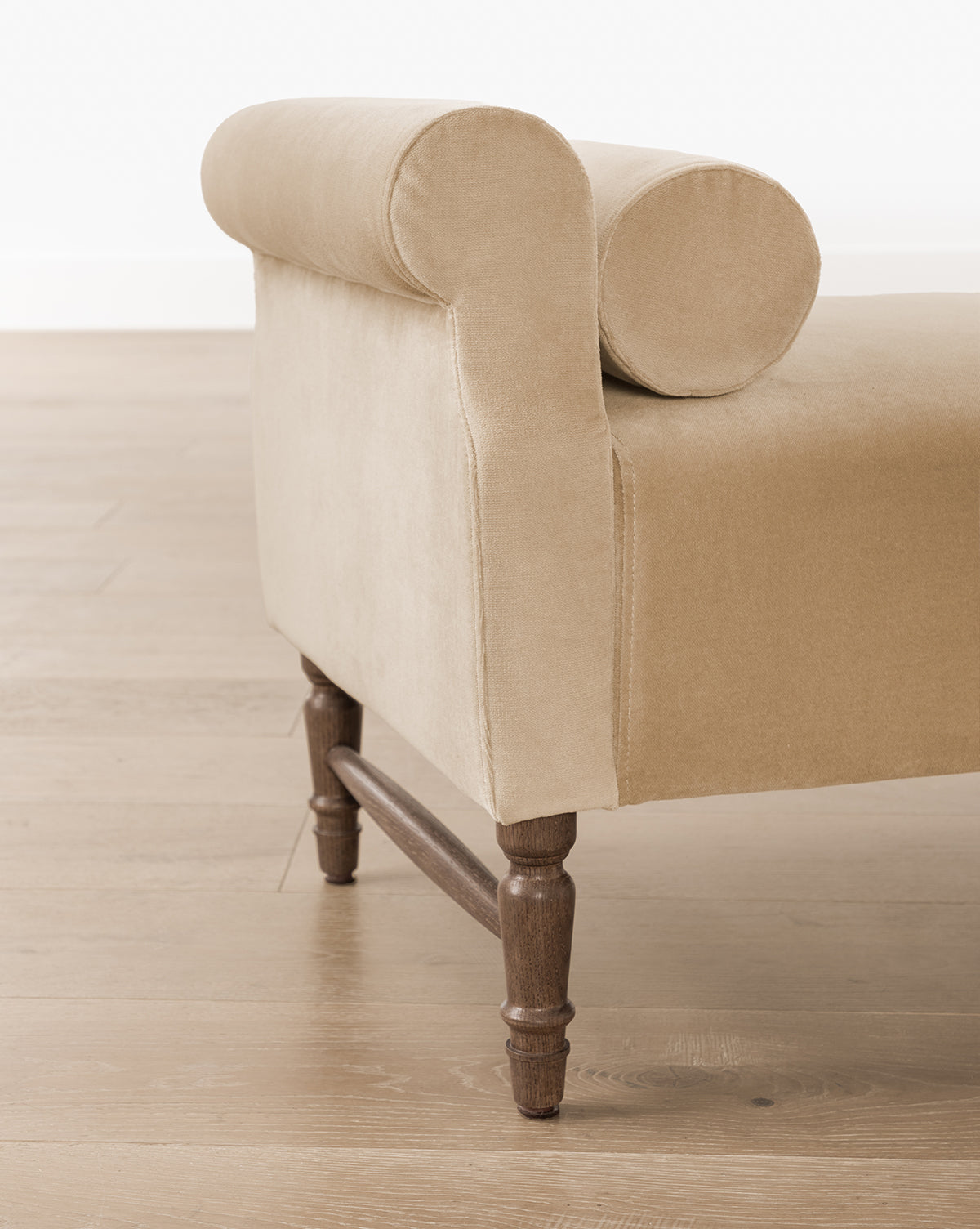 The Asher Bench is a beige upholstered chaise lounge with a rolled armrest, round cushion, bolster pillows, and wooden legs, shown on a light wooden floor, by Vendixs