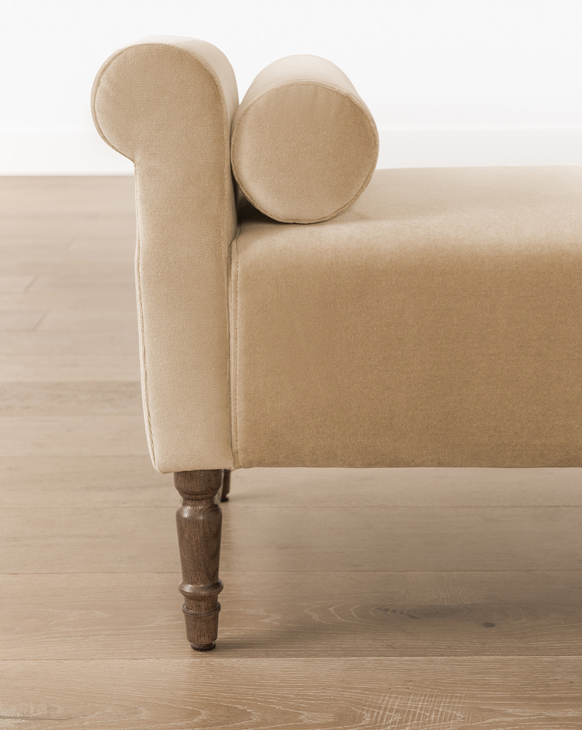 The Asher Bench, featuring beige striped linen upholstery and bolster pillows, rests on turned wooden legs atop a light wood floor with a white wall backdrop, by Vendixs