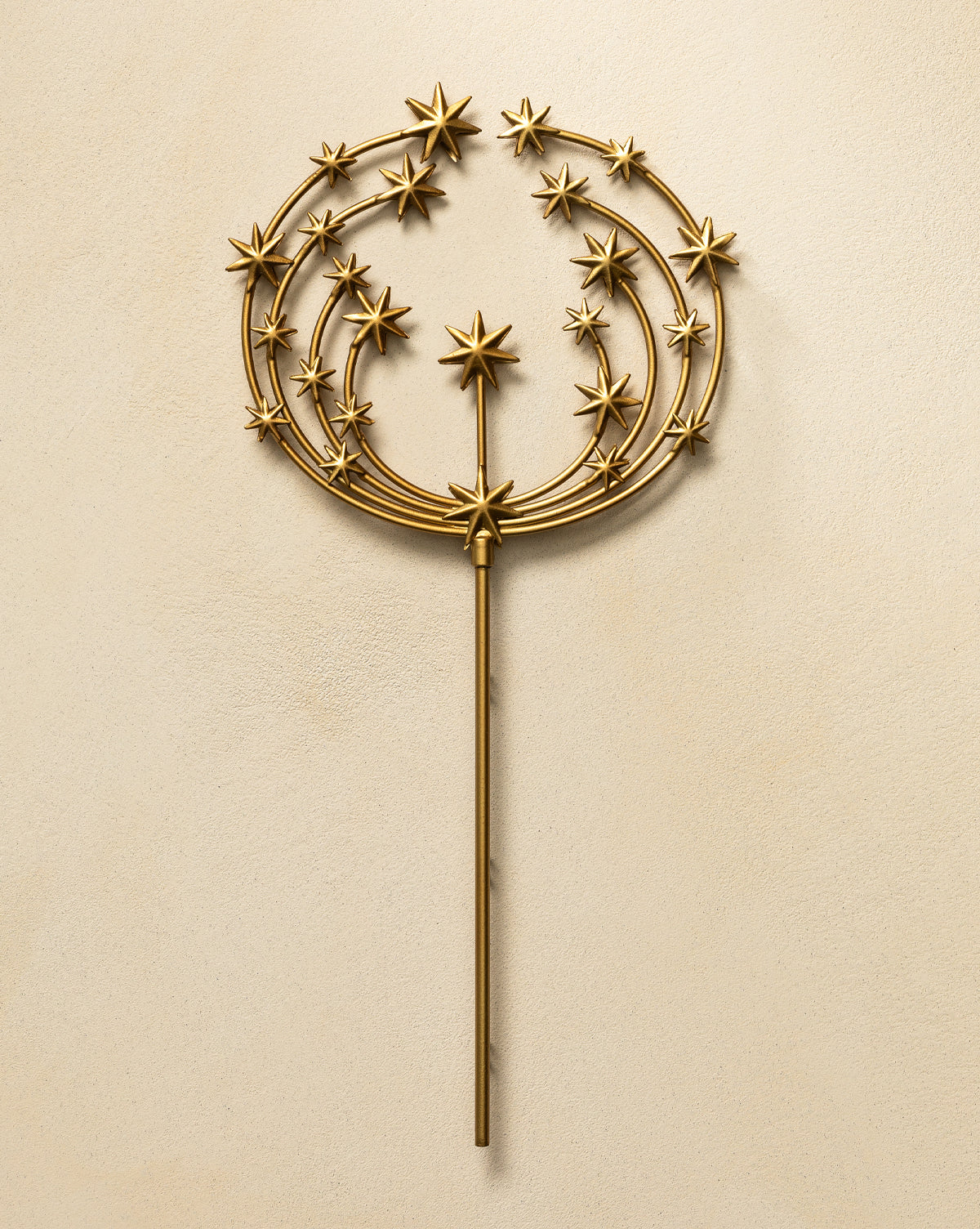 The Astra Brass Tree Topper features a gold metal wand with an antique brass finish, a round top adorned with stars, and three concentric arcs radiating from a central star, set on a beige textured background—ideal for holiday decor, by Vendixs