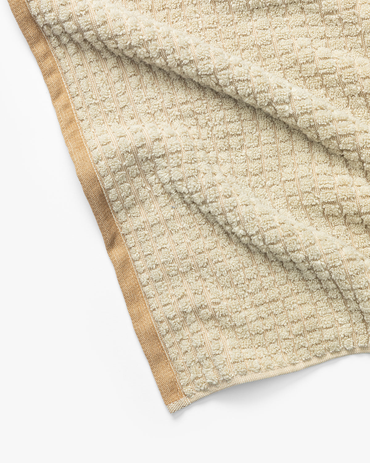 A close-up of a beige textured waffle weave towel from the Atrani Organic Bath Collection, crafted from organic cotton with a solid tan border, laid out on a white surface, by Vendixs