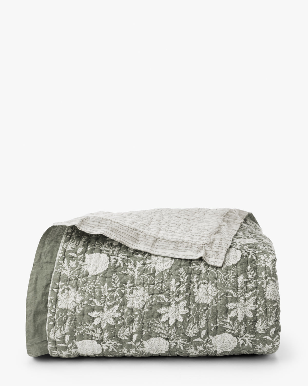The Audrey Sage Block Print Quilt, featuring a green and white floral pattern with subtle ruffle edging, is folded and topped with a beige textured blanket against a plain white background, by Vendixs
