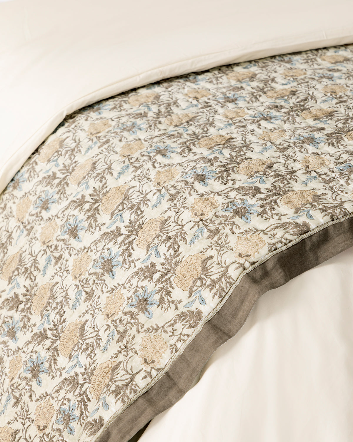 A close-up of a bed with the Audrey Tan Block Print Quilt, showcasing beige, brown, and light blue floral patterns on a white background with a gray border, layered over a plain white duvet, by Vendixs