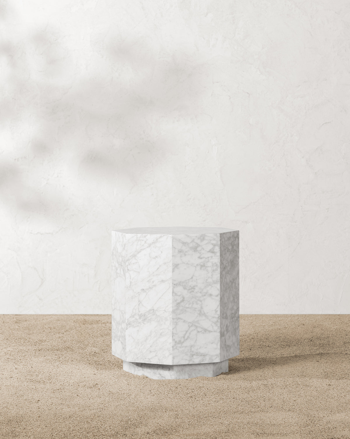 The Barcelo Outdoor Side Table, crafted from white marble with subtle grey veining, features a geometric design and stands on a sandy surface against a textured off-white wall, by Vendixs