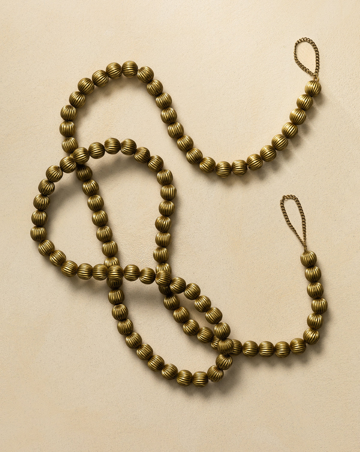 The 6' Batavia Brass Garland features round, gold-colored, ribbed beads with an antique brass finish and a looped end, arranged on a light beige surface—perfect for adding festive holiday decor to your home, by Vendixs