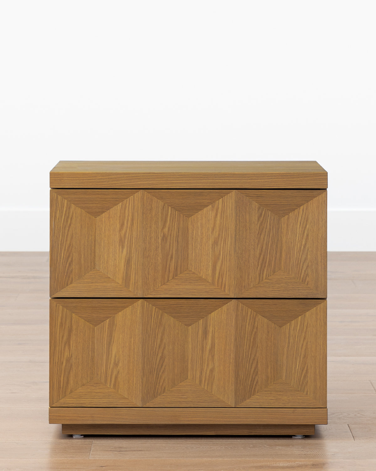The Beacon Nightstand, made from white oak, features two drawers with a geometric textured front, offering stylish bedroom storage, by McGee & Co. It sits on a wooden floor against a plain white wall for a modern look.