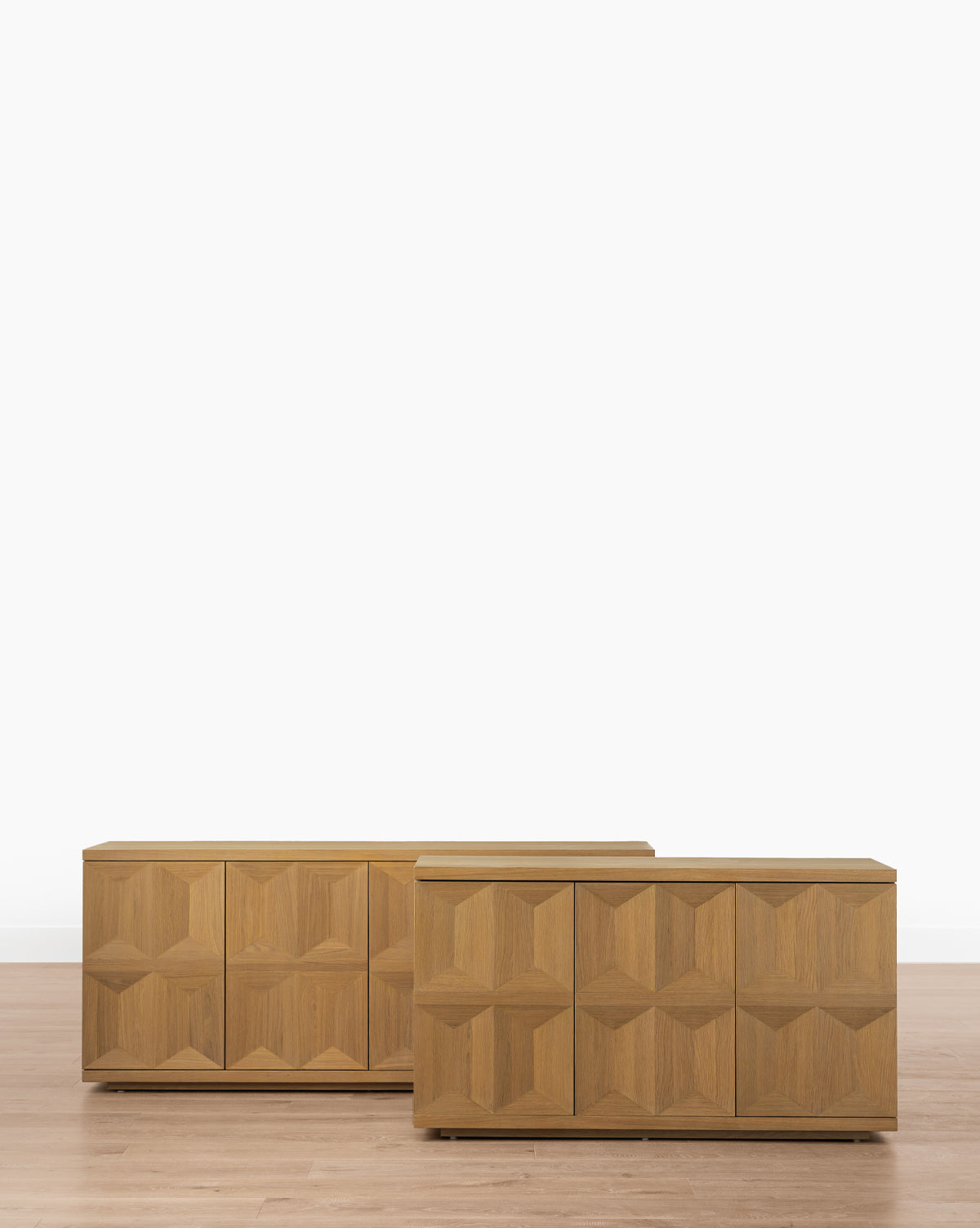 Two Beacon Sideboards with geometric, textured fronts and a neutral finish stand on a light wood floor by a white wall. One is slightly smaller and placed in front of the other, providing stylish and spacious storage by Vendixs