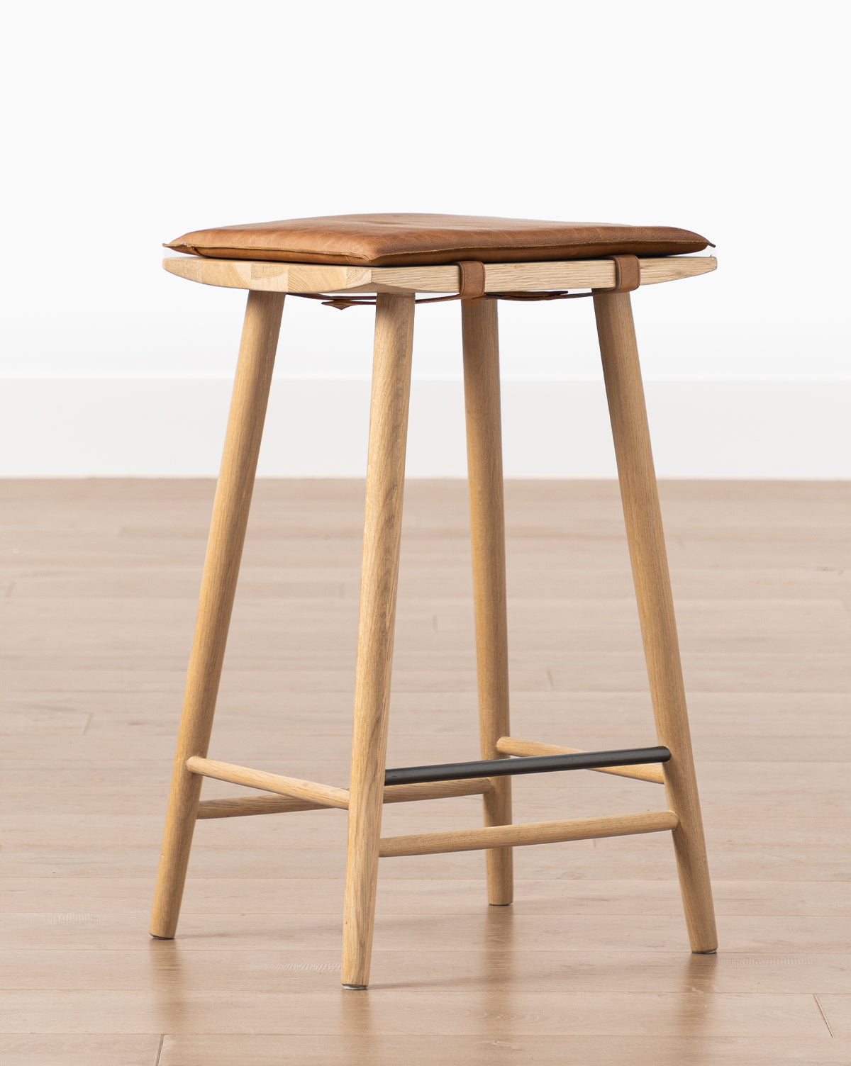 The Beau Counter Stool features a modern design with a wooden frame, four angled legs, a square brown leather seat cushion, and a black metal footrest, by Vendixs