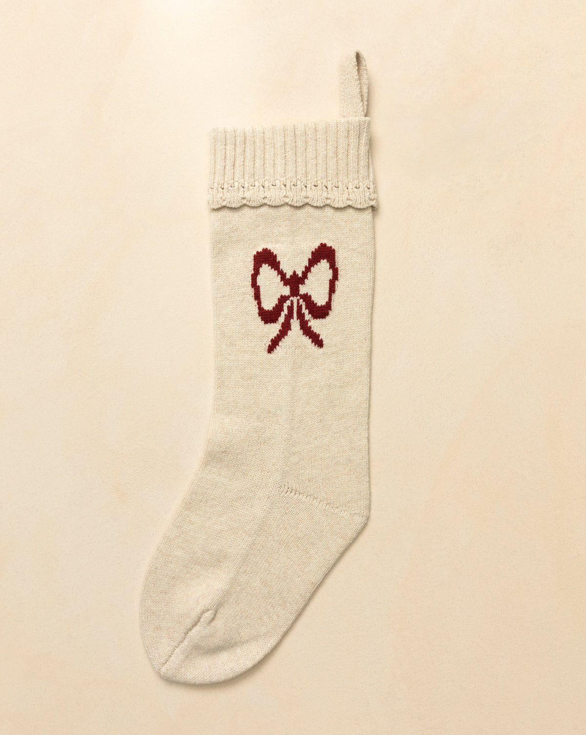 The Bella Scalloped Stocking with Bow is a cream knit Christmas stocking featuring a ribbed cuff, hanging loop, and red bow on the front—ideal festive mantel decor—shown on a neutral background, by Vendixs
