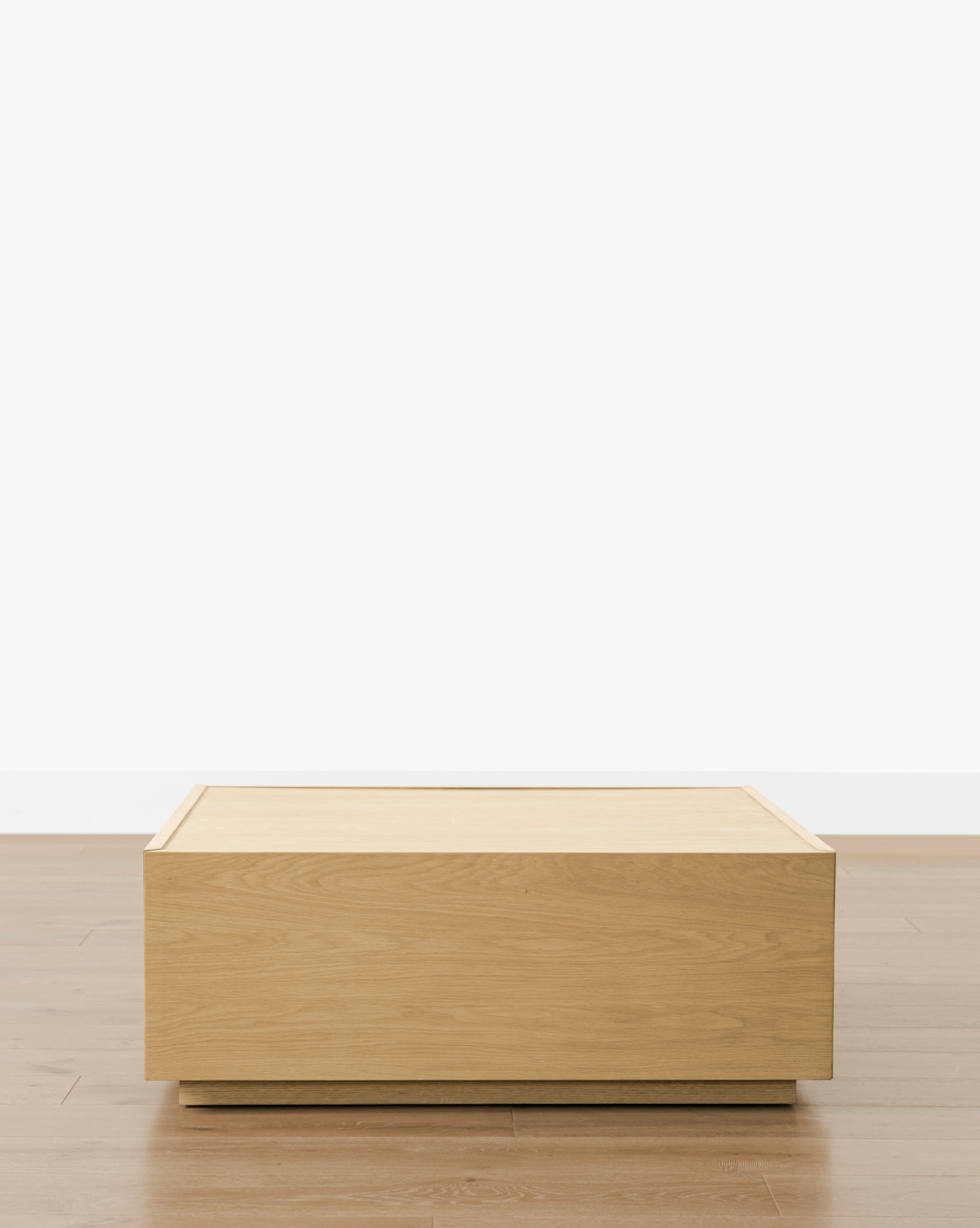 The Bentley Coffee Table stands on a light brown wood floor against a plain white wall, offering a clean, minimalist look, by Vendixs
