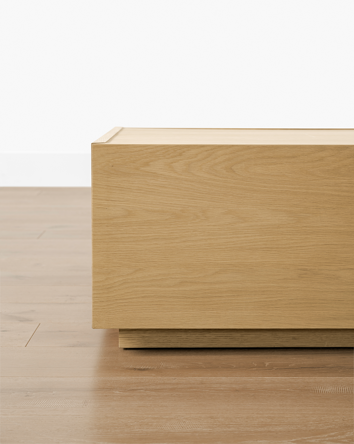 A close-up of the Bentley Coffee Table, featuring a rectangular, light oak finish. Positioned on a wooden floor by a white wall, only its stylish corner is visible, by Vendixs