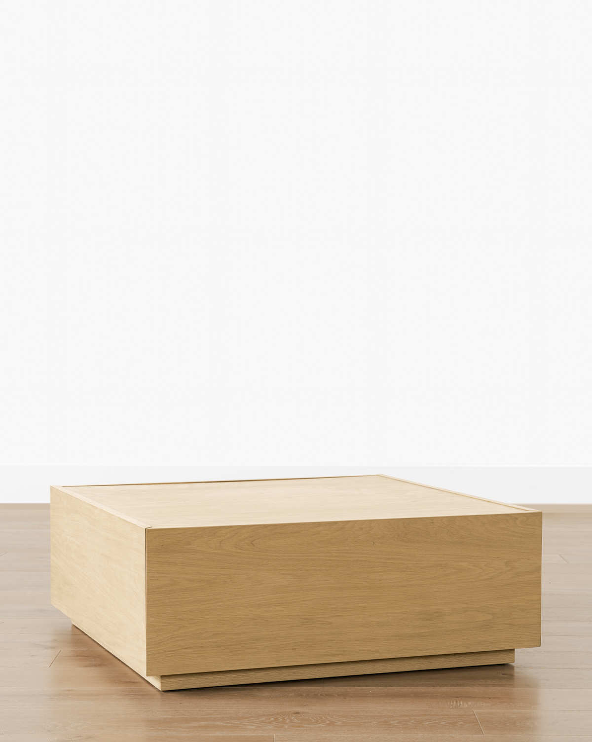 The Bentley Coffee Table is a minimalist, square-shaped design in light oak, featuring clean lines and a solid, box-like look. Perfect for modern spaces, it sits beautifully on wooden floors against plain white walls, by Vendixs