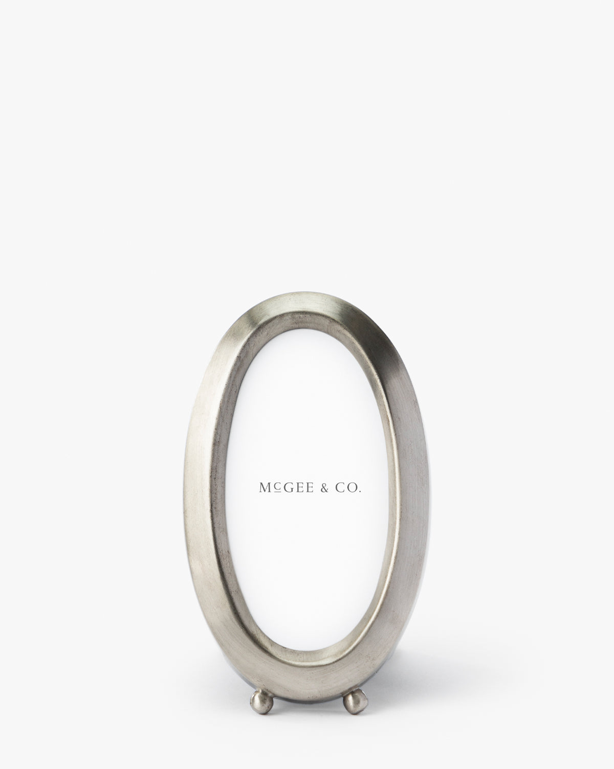 The Blanche Pewter Picture Frame features a sleek oval silver design with a modern, sophisticated style. Standing upright on two small round feet, it displays a MEGEE & CO. placeholder card against a plain white background.