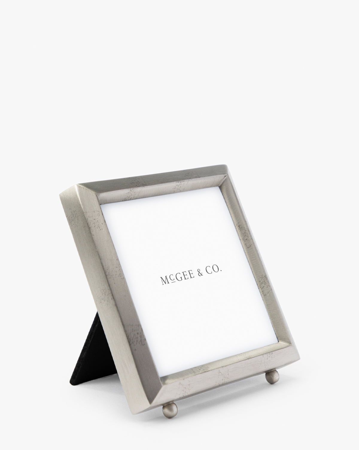The Blanche Pewter Picture Frame, a square metallic design with iron corners and stand, holds an Vendixs insert and exudes sophistication as it stands angled against a plain white background.