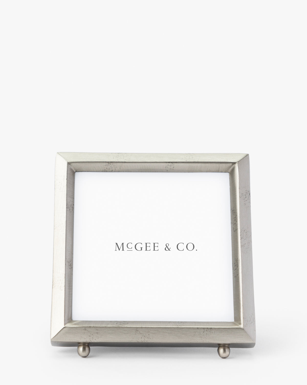 The Blanche Pewter Picture Frame is a square, silver metal frame with a simple modern design, iron corners, and two small round feet. It features a white placeholder displaying Vendixs