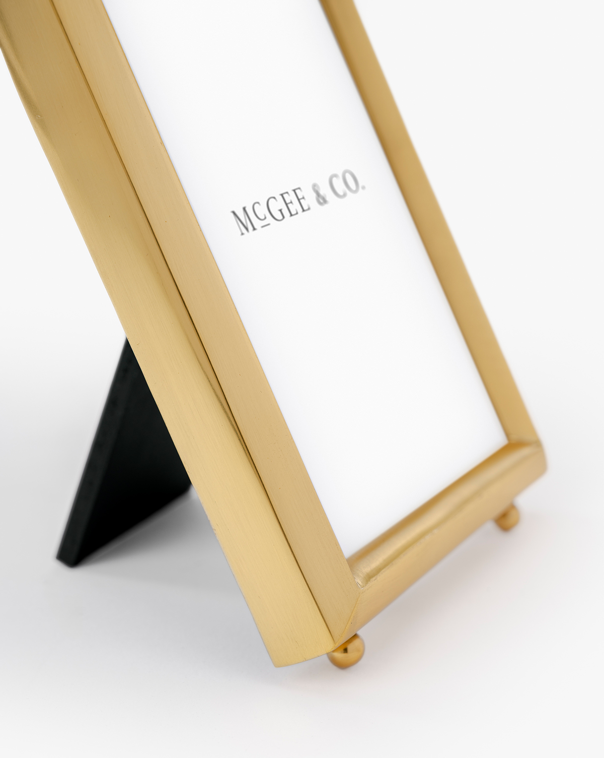 A close-up of the Blanche Picture Frame in polished brass stands on a black support, displaying "McGee & Co." on the paper inside, set against a plain white background, by Vendixs