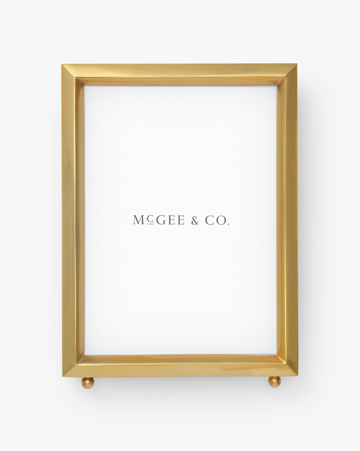 The Blanche Picture Frame is a rectangular gold frame with a white background, featuring "MCGEE & CO." centered. Its modern, elegant design sits on two small gold spheres at the base, by Vendixs
